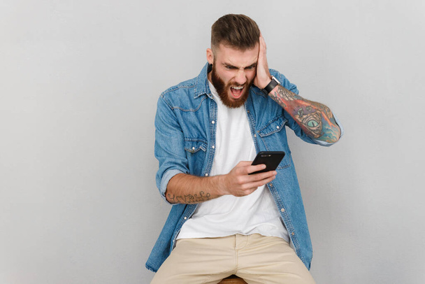 Portrait of a handsome angry young man wearing casual clothes sitting isolated over gray background, screaming at his mobile phone - Fotó, kép