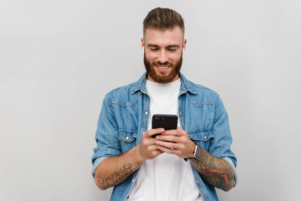 Portrait of a handsome smiling young man wearing casual clothes standing isolated over gray background, listening to music with wireless earphones, holding mobile phone - Valokuva, kuva