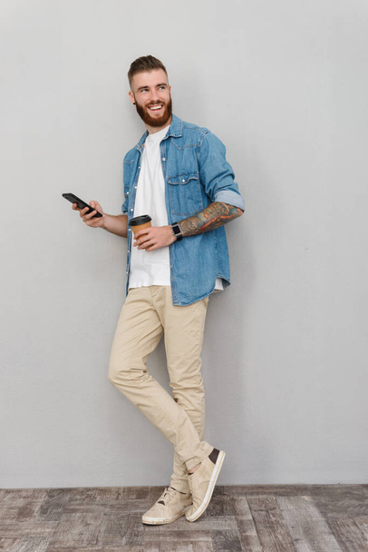 Full length portrait of a handsome smiling young man wearing casual clothes standing over gray background, using mobile phone while drinking takeaway coffee cup - Fotó, kép