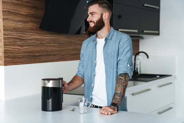 Smiling young bearded man making cup of tea while standing in the kitchen at home - Φωτογραφία, εικόνα