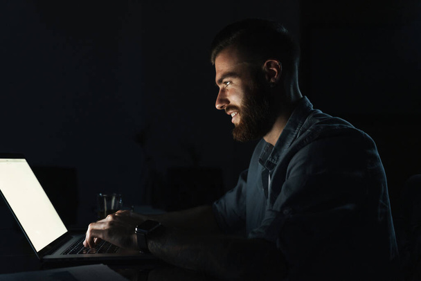 Un jeune homme confiant et souriant qui travaille sur un ordinateur portable assis à la table à l'intérieur la nuit
 - Photo, image