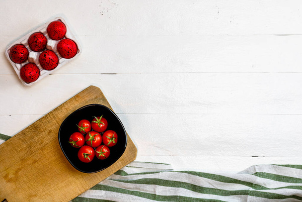 Vue de dessus des tomates cerises fraîches mûres et des œufs de Pâques rouges sur une table rustique en bois blanc. Ingrédients et concept alimentaire - Photo, image