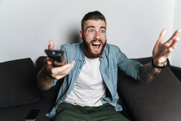 Attractive excited young bearded man relaxing on a couch at home, watching tv, screaming - Foto, immagini