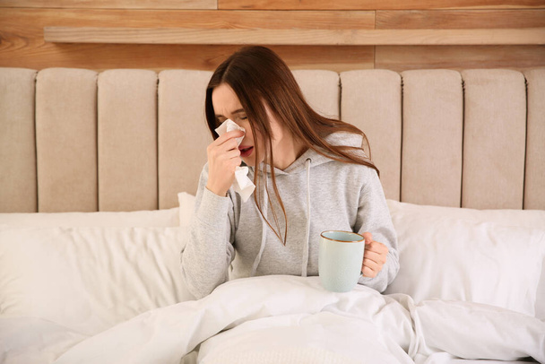 Sick young woman with cup of hot drink in bed at home. Influenza virus - Фото, изображение