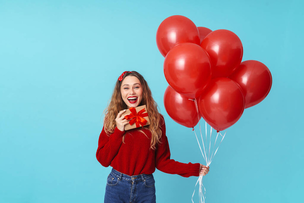 Image of happy blonde woman laughing while holding gift box and balloons isolated over blue background - Foto, Imagem