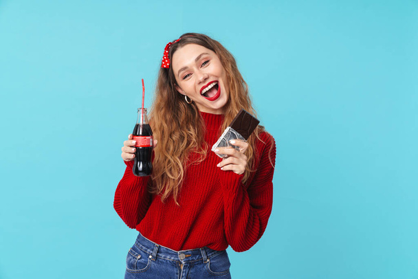Image of smiling pretty woman drinking soda and eating chocolate isolated over blue background - Fotografie, Obrázek