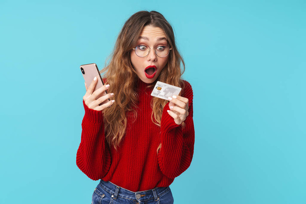 Image of surprised caucasian woman in eyeglasses holding cellphone and credit card isolated over blue background - Φωτογραφία, εικόνα