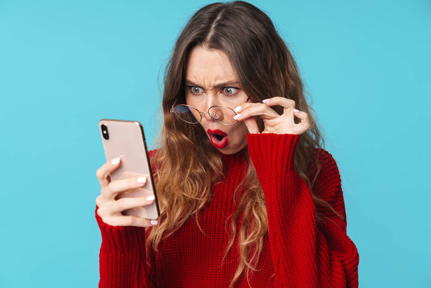 Εικόνα δυσαρεστημένης καλής γυναίκας με γυαλιά ηλίου χρησιμοποιώντας κινητό τηλέφωνο και εκφράζοντας έκπληξη απομονωμένη σε μπλε φόντο - Φωτογραφία, εικόνα