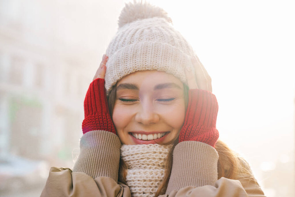 Close up of a cheerful pretty young girl wearing winter jacket and a hat standing on a city street - Photo, Image