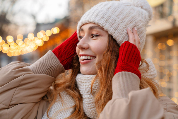 Close up of a cheerful pretty young girl wearing winter jacket and a hat standing on a city street - Foto, Bild