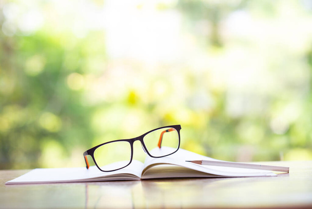 Black eyeglasses, white pencil with white notebook on wooden table, Bokeh garden background, Close up & Macro shot, Selective focus, Stationery concept - Φωτογραφία, εικόνα