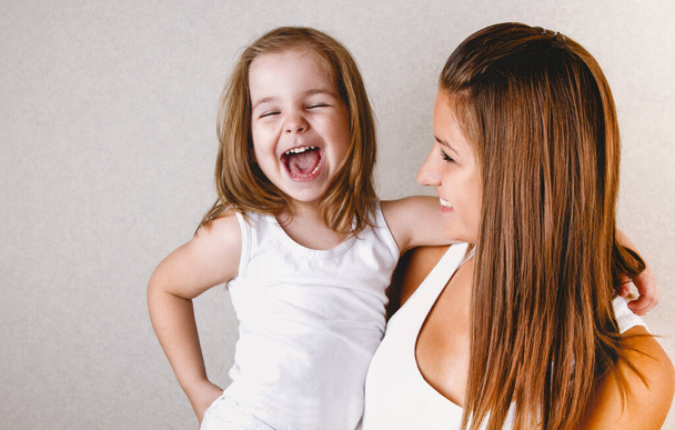 Sonriente joven pelirroja casual hembra sosteniendo riéndose hija pequeña en camiseta blanca mientras se divierten juntos de pie contra fondo claro
 - Foto, Imagen