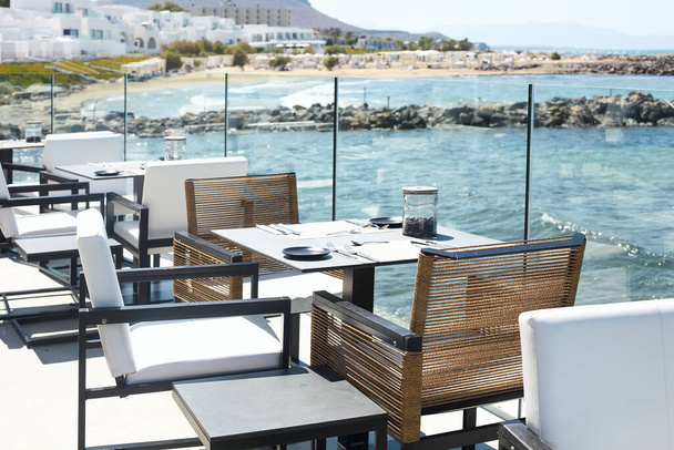 Sillas y mesas con vajilla situada cerca de la valla de vidrio en la terraza del restaurante de lujo cerca del mar ondulante en el día soleado en el complejo
 - Foto, Imagen