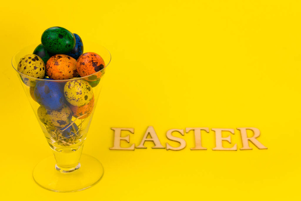 Multi-colored Easter eggs in a glass cup and the inscription Easter on a yellow background. - Photo, Image