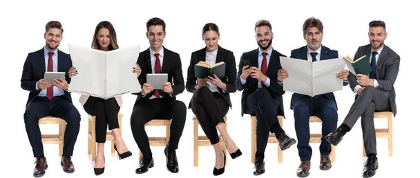 Team of 7 businessmen reading from books, newspaper, tables and phones while waiting for a job interview, sitting on chairs on white studio background - Photo, Image