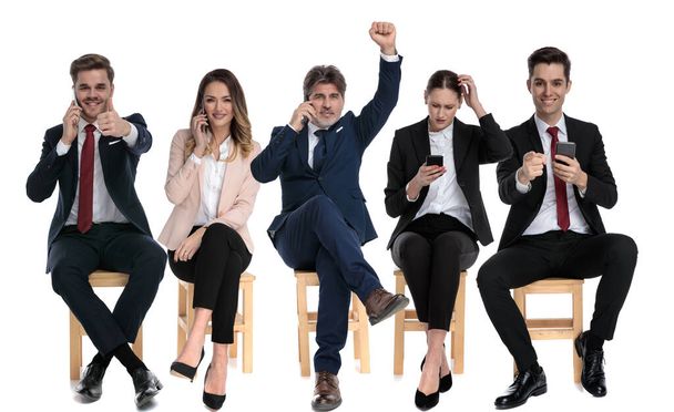 Team of 5 businessmen talking, reading and writing on their phones, celebrating while waiting for a job interview sitting on chairs on white studio background - Photo, Image