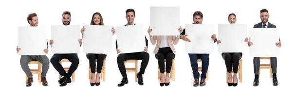 Team of 8 businessmen holding empty billboards while one is hiding her face while sitting on chairs on white studio background - Photo, Image