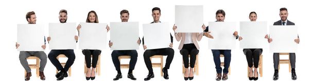Equipo de 9 hombres de negocios sosteniendo vallas publicitarias vacías mientras uno está escondiendo su cara, sentado en sillas sobre fondo blanco del estudio
 - Foto, imagen