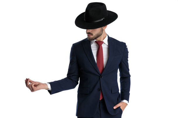 Şapka ve takım elbise giyerken elini cebinde şaklatan gizemli bir adam beyaz stüdyo arka planında duruyor. - Fotoğraf, Görsel