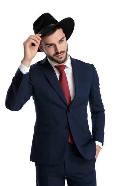 Encantador empresário cumprimentando educadamente com a mão no bolso enquanto usava chapéu e terno, de pé no fundo do estúdio branco
 - Foto, Imagem