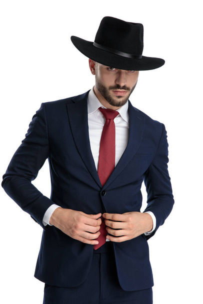 Sério olhando empresário fixando seu terno enquanto vestindo chapéu e terno, de pé sobre fundo estúdio branco
 - Foto, Imagem