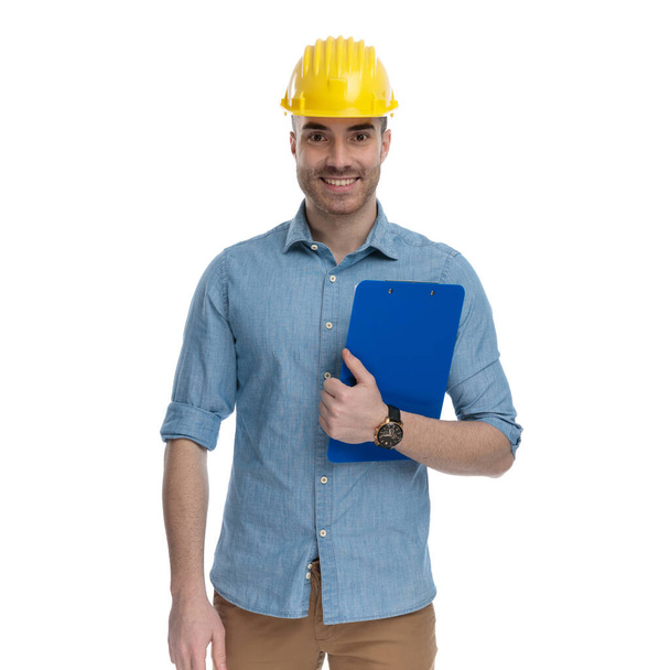 Happy casual man holding clipboard and wearing safety helmet, standing on white studio background - Zdjęcie, obraz