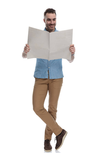 Glücklicher, lässiger Mann, der Zeitung liest und lächelt, während er blaues Hemd auf weißem Studiohintergrund trägt - Foto, Bild
