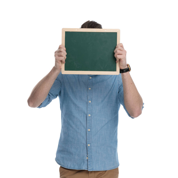 Jeune homme décontracté couvrant son visage avec un tableau vert tout en portant une chemise bleue, debout sur fond de studio blanc
 - Photo, image