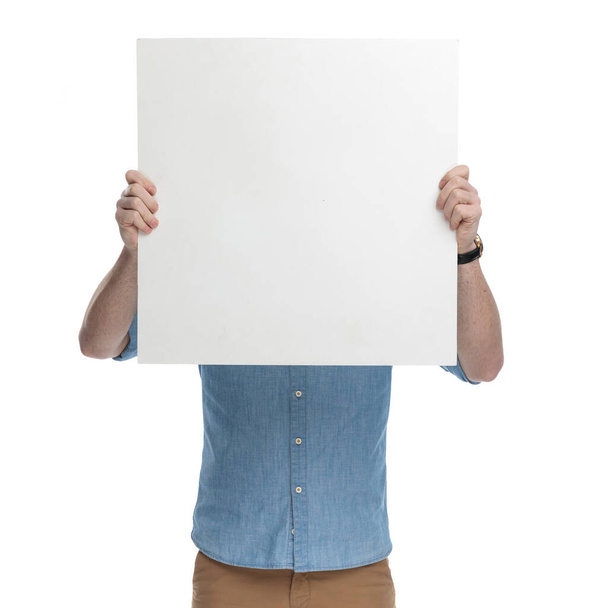 Młody mężczyzna zakrywający twarz czystym billboardem w niebieskiej koszuli, stojący na białym tle studia - Zdjęcie, obraz