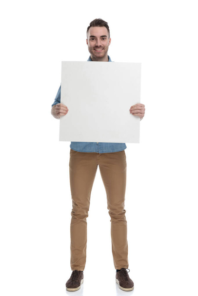 Šťastný ležérní muž se usmívá a drží prázdný billboard při nošení modré košile, stojí na bílém pozadí studia - Fotografie, Obrázek