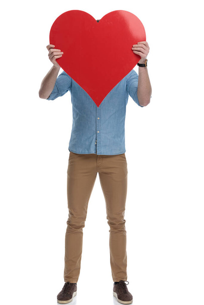 Young casual man covering his face with a heart shape while wearing blue shirt, standing on white studio background - Foto, Imagen