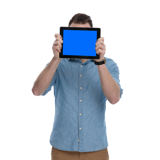 Mavi gömlek giyerken yüzünü boş tabletle örten genç bir adam beyaz stüdyo arka planında duruyor. - Fotoğraf, Görsel