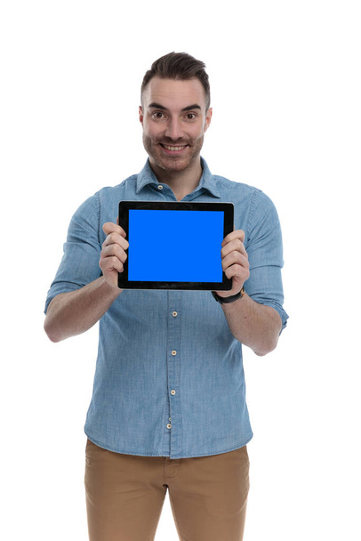 Happy casual man smiling and presenting empty tablet while wearing blue shirt, standing on white studio background - Foto, Imagen