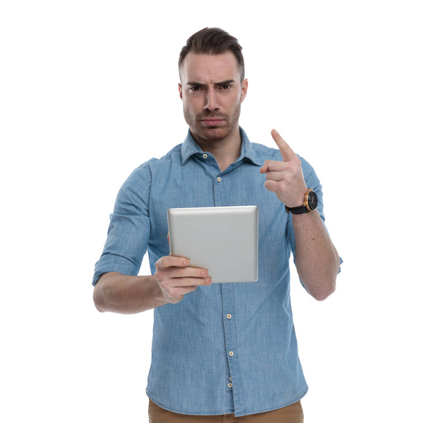 Molesto hombre casual sosteniendo la tableta y culpando mientras usa camisa azul, de pie sobre fondo de estudio blanco
 - Foto, imagen