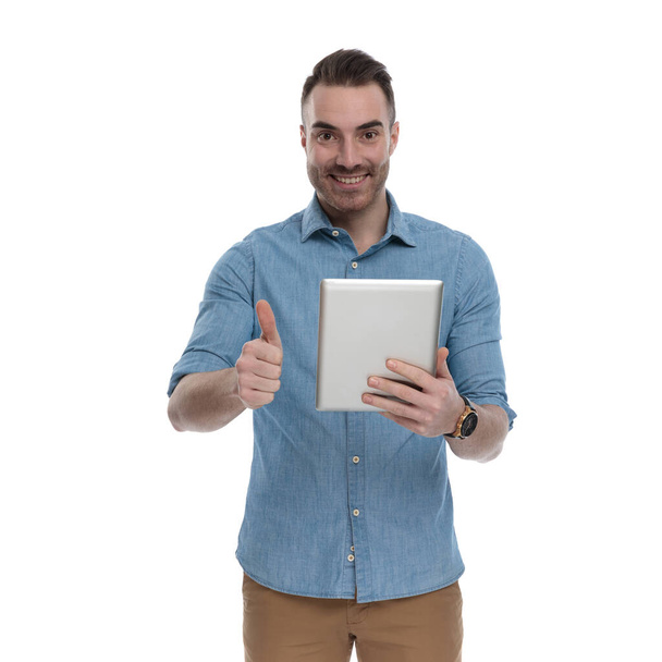 Positiv lässiger Mann mit Tablette, lächelnd und gestikulierend, während er blaues Hemd trägt, auf weißem Studiohintergrund stehend - Foto, Bild
