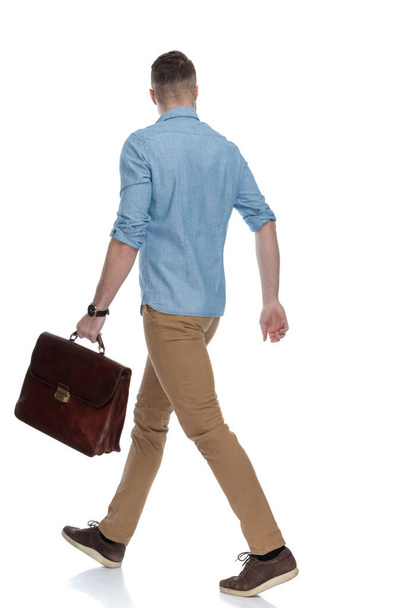 Side view of eager casual holding briefcase while wearing blue shirt, walking on white studio background - Fotografie, Obrázek