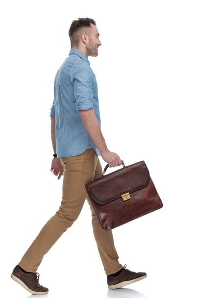 Side view of positive casual holding briefcase and smiling while wearing blue shirt, stepping on white studio background - Foto, imagen
