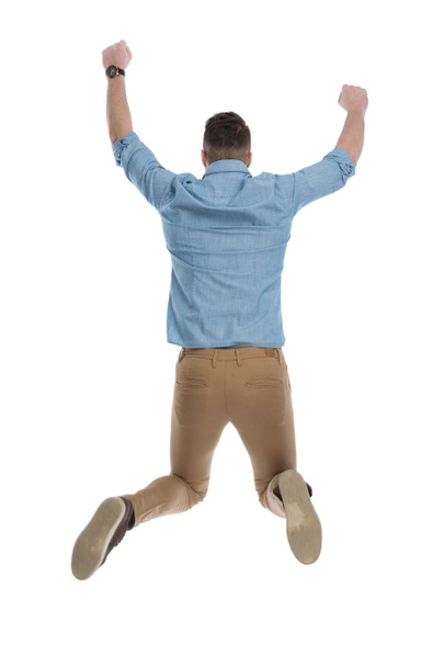 Rear view of celebrating casual man jumping while wearing blue shirt on white studio background - Photo, Image