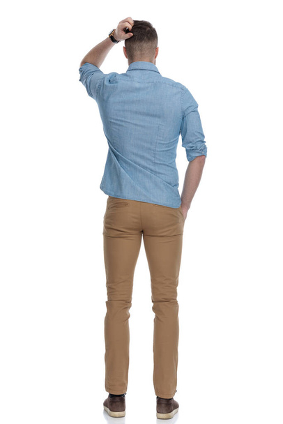 Rear view of confused casual man scratching his head while wearing blue shirt, standing on white studio background - Photo, Image