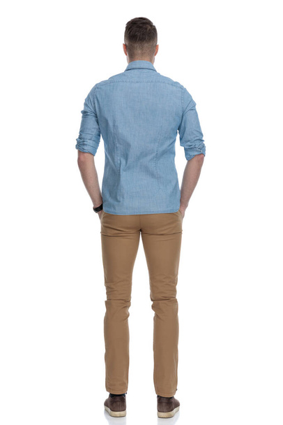 白いスタジオの背景に立って、青いシャツを着ている間、彼のポケットに彼の手を保持カジュアル男の背面図 - 写真・画像