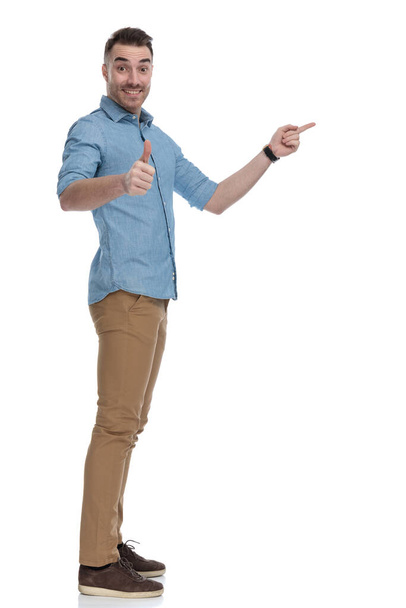 Vista lateral del alegre hombre casual señalando y haciendo gestos bien mientras usa camisa azul, de pie sobre fondo blanco del estudio
 - Foto, imagen