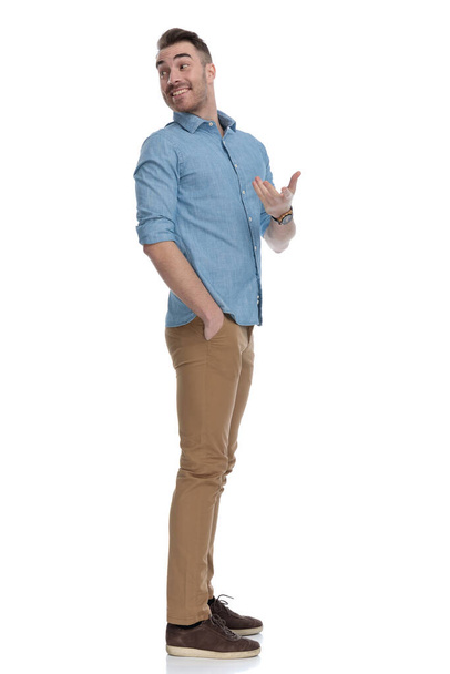 青いシャツを着て笑って説明する陽気なカジュアルな男のサイドビュー、白いスタジオの背景に立って - 写真・画像