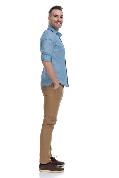 Side view of happy  casual man smiling with both hands in pockets while wearing blue shirt, standing on white studio background - Fotoğraf, Görsel
