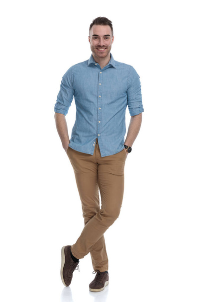 Gelukkig casual man glimlachen met beide handen in zijn zakken terwijl het dragen van een blauw shirt, staan op witte studio achtergrond - Foto, afbeelding