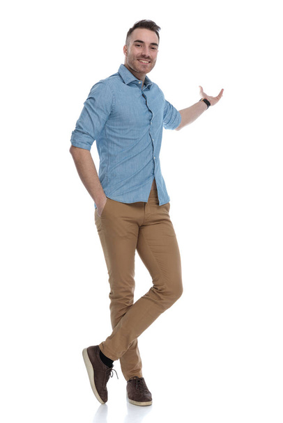 Charming casual man presenting with his hand in his pocket, smiling while wearing blue shirt, standing on white studio background - Photo, Image
