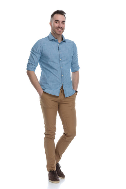 Positive casual man smiling with both hand in his pockets while wearing blue shirt, standing on white studio background - 写真・画像
