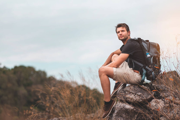 Portret aktywnego brodatego podróżnika w czarnej koszuli siedzącego i cieszącego się zapierającą dech w piersiach przyrodą scenerią wzgórz na szczycie gór. Szczęśliwy relaksujący mężczyzna z plecakiem patrząc na piękny krajobraz. Turysta lub alpinista spoczywający na szczycie. - Zdjęcie, obraz