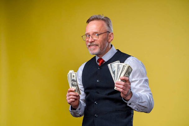 Happy exited man in glasses and white shirt holding bunch of money, looking at camera, isolated over yellow background - Photo, image