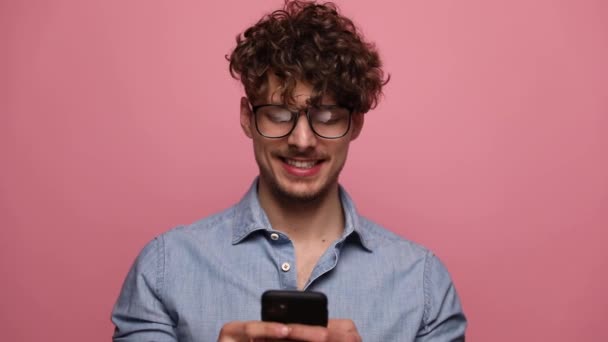 atrakcyjny dorywczy mężczyzna nosi okulary stojąc i trzymając telefon szczęśliwy następnie podnosząc jedną pięść podekscytowany na różowym tle studio - Materiał filmowy, wideo