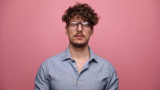 vonzó alkalmi férfi visel szemüveg áll, és nézi a kamera komoly, majd tegye a kezét az arcára meglepett rózsaszín stúdió háttér - Felvétel, videó